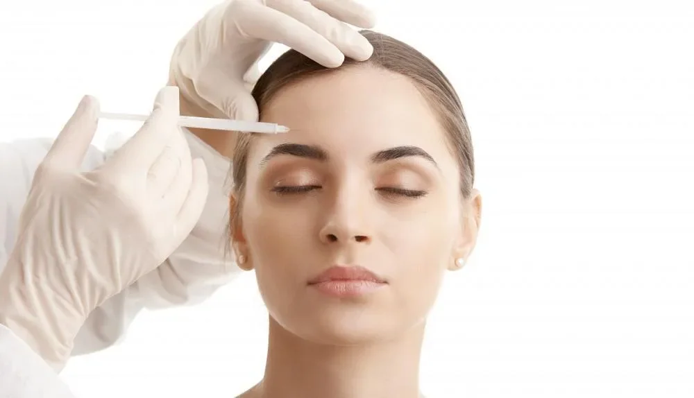 Gloved hands positioning a cosmetic injection near a woman’s forehead